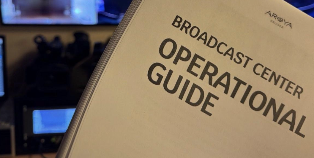 Broadcast Center Operational Guide held in front of a multi-view monitor wall